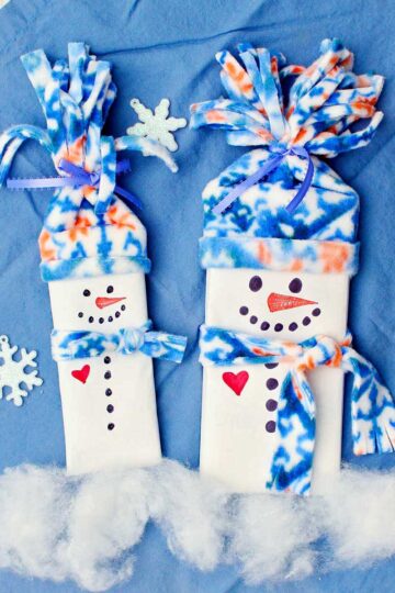 Two completed candy bar snowman stocking stuffers with fleece hats and scarves against a blue cloth with sparkly snowflake decorations.