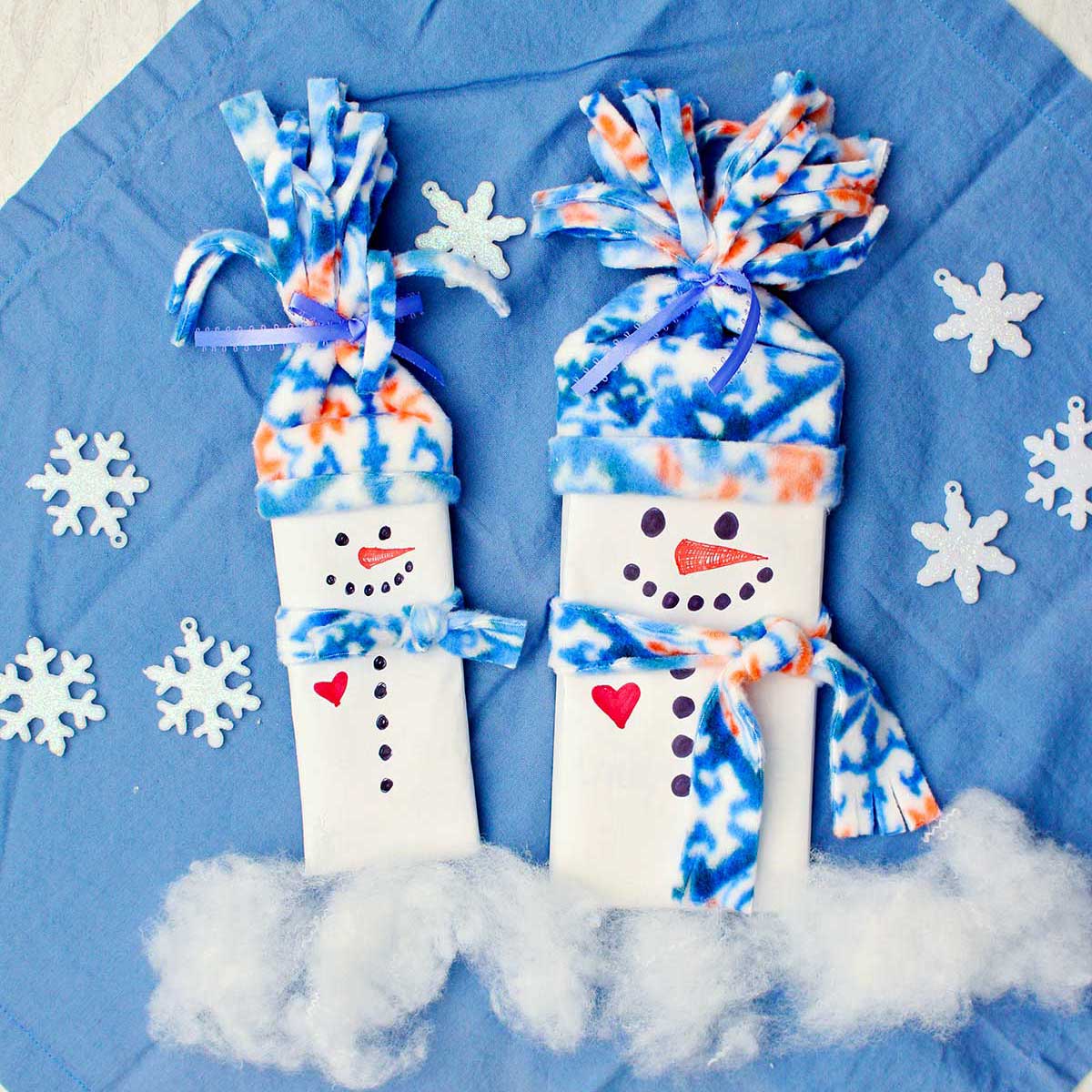 Two completed candy bar snowman stocking stuffers with fleece hats and scarves against a blue cloth with sparkly snowflake decorations.