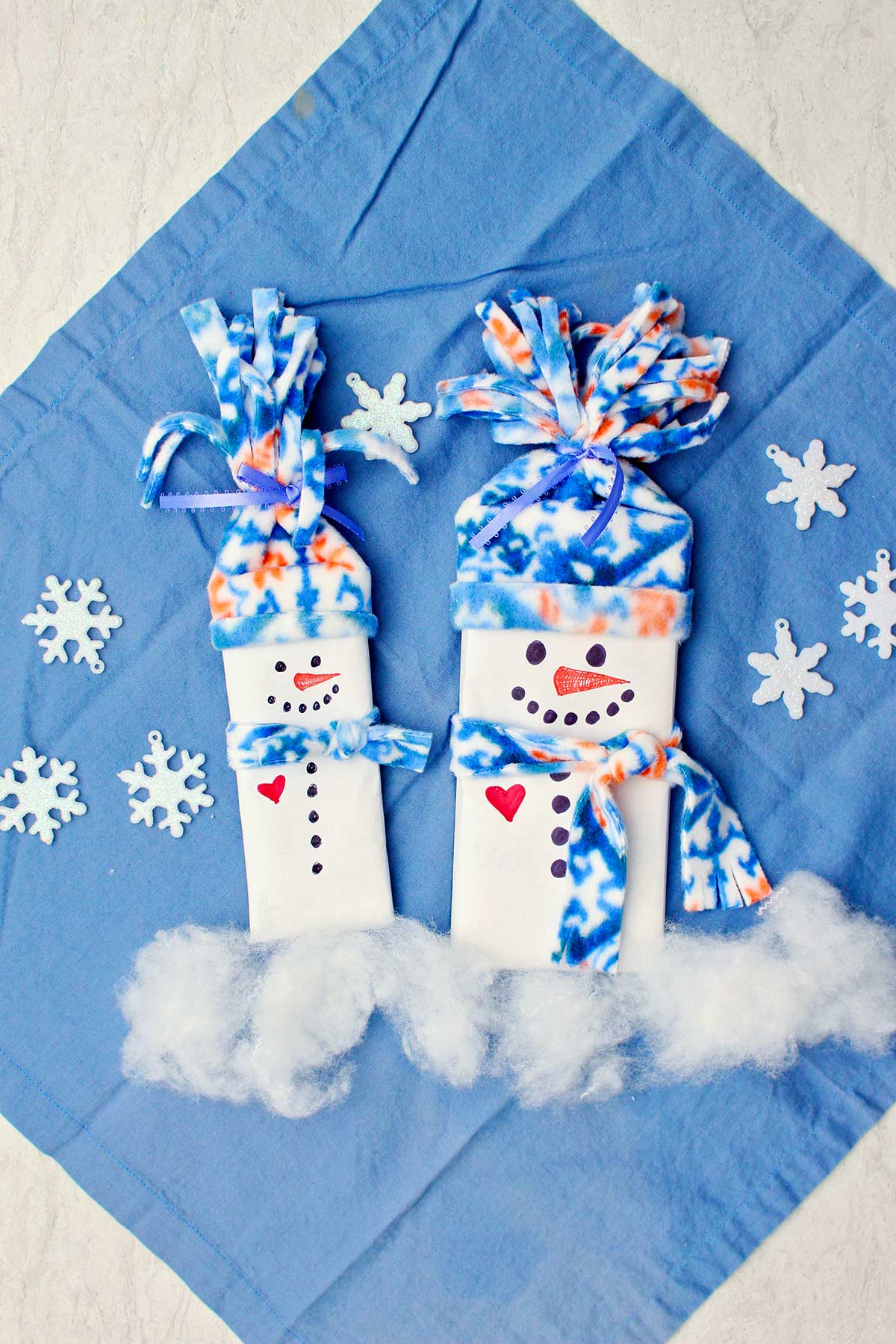 Two completed candy bar snowman stocking stuffers with fleece hats and scarves against a blue cloth with sparkly snowflake decorations.