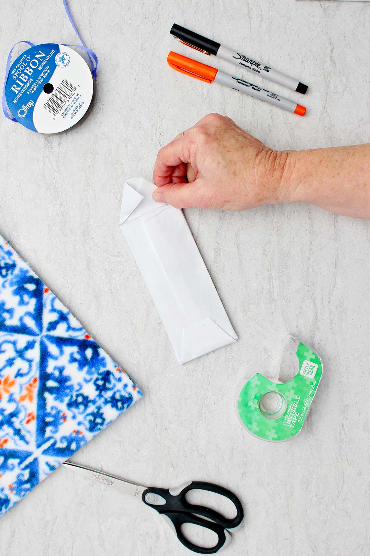 Person folding down last bits of white paper to cover candy bar in white paper with other supplies to make snowman stocking stuffer near by.