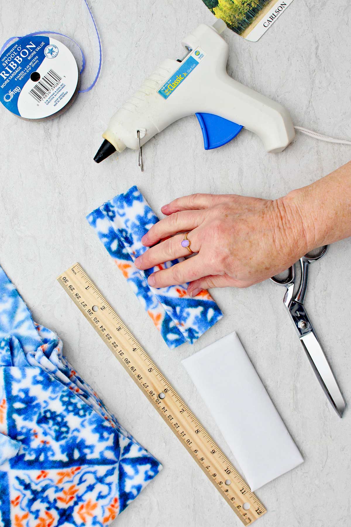 Person folding blue and orange patterned fleece and hot gluing edges to make hat for snowman stocking stuffer with other supplies to make snowman stocking stuffer near by.