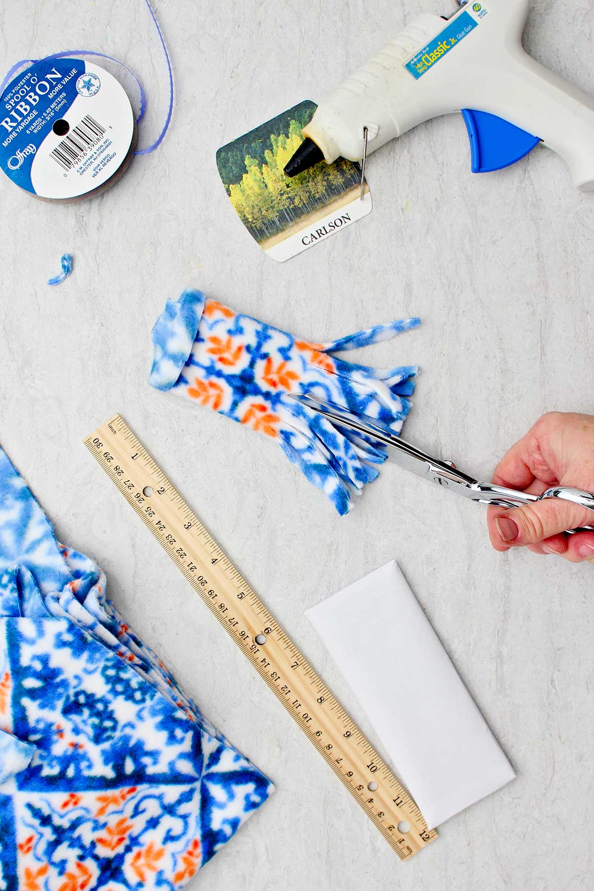 Person cutting fringe in blue and orange patterned fleece with other supplies to make snowman stocking stuffer near by.