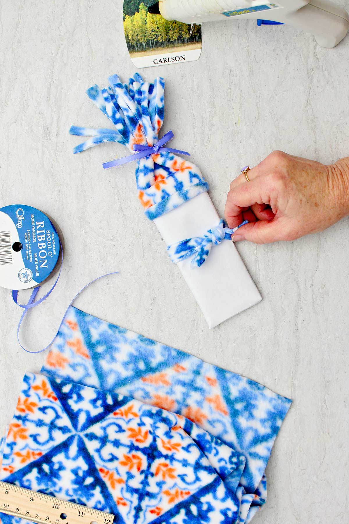 Person securing piece of fleece around the middle of the wrapped candy bar to make a scarf with other supplies to make snowman stocking stuffer near by.