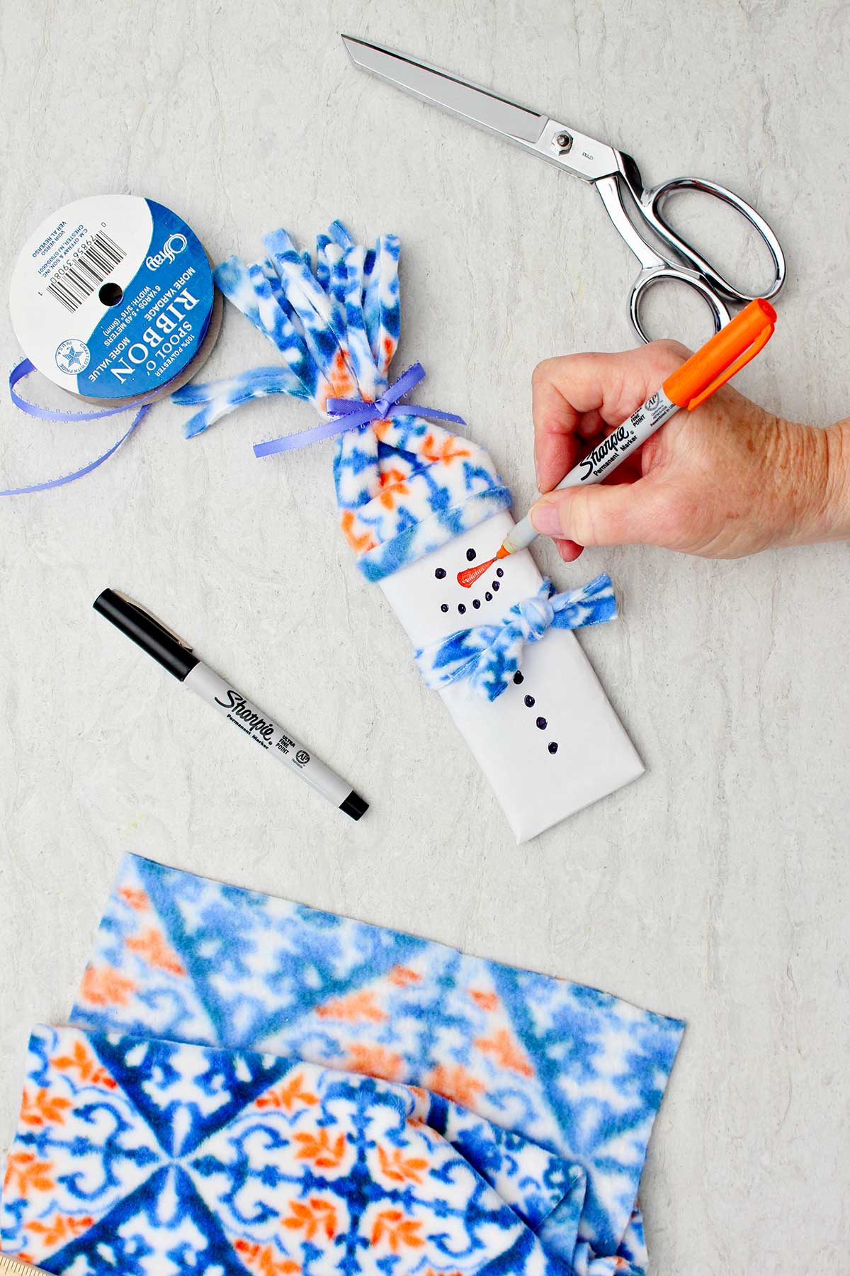 Person drawing a carrot nose with sharpie on the white paper wrapped candy bar with other supplies to make snowman stocking stuffer near by.