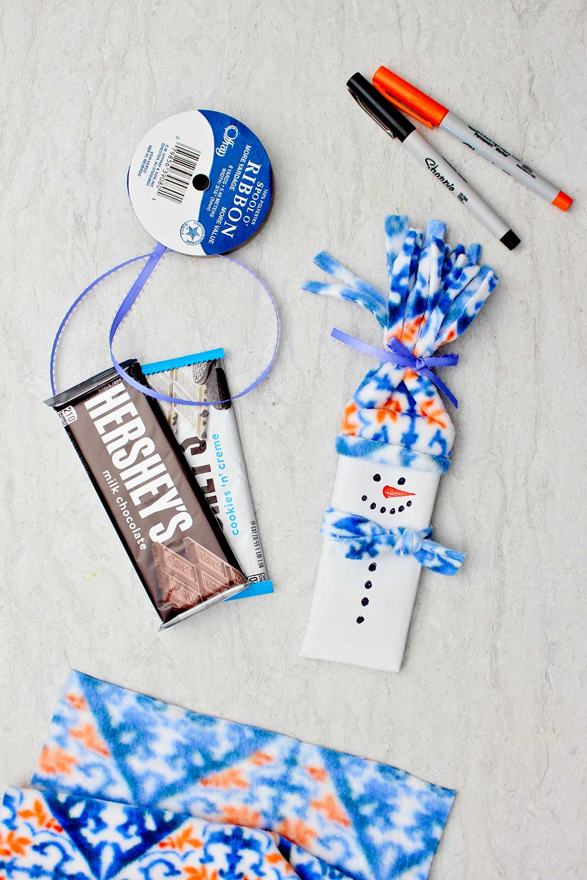 Completed candy bar snowman stocking stuffer with candy bars, fleece, ribbon and sharpies near by.