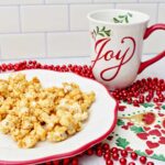 Caramel corn on a white plate with a red rim, a holiday napkin, red beads and a holiday mug that says "joy".