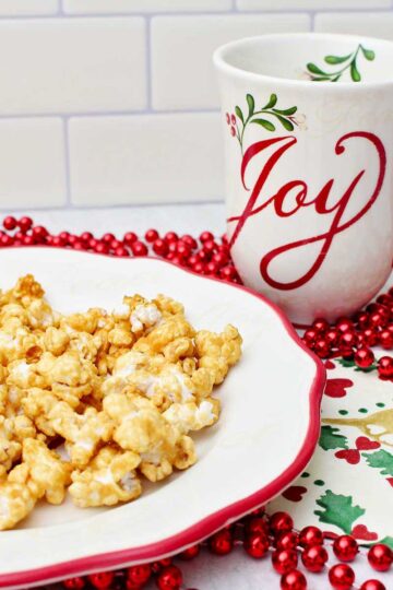 Caramel corn on a white plate with a red rim, a holiday napkin, red beads and a holiday mug that says "joy".