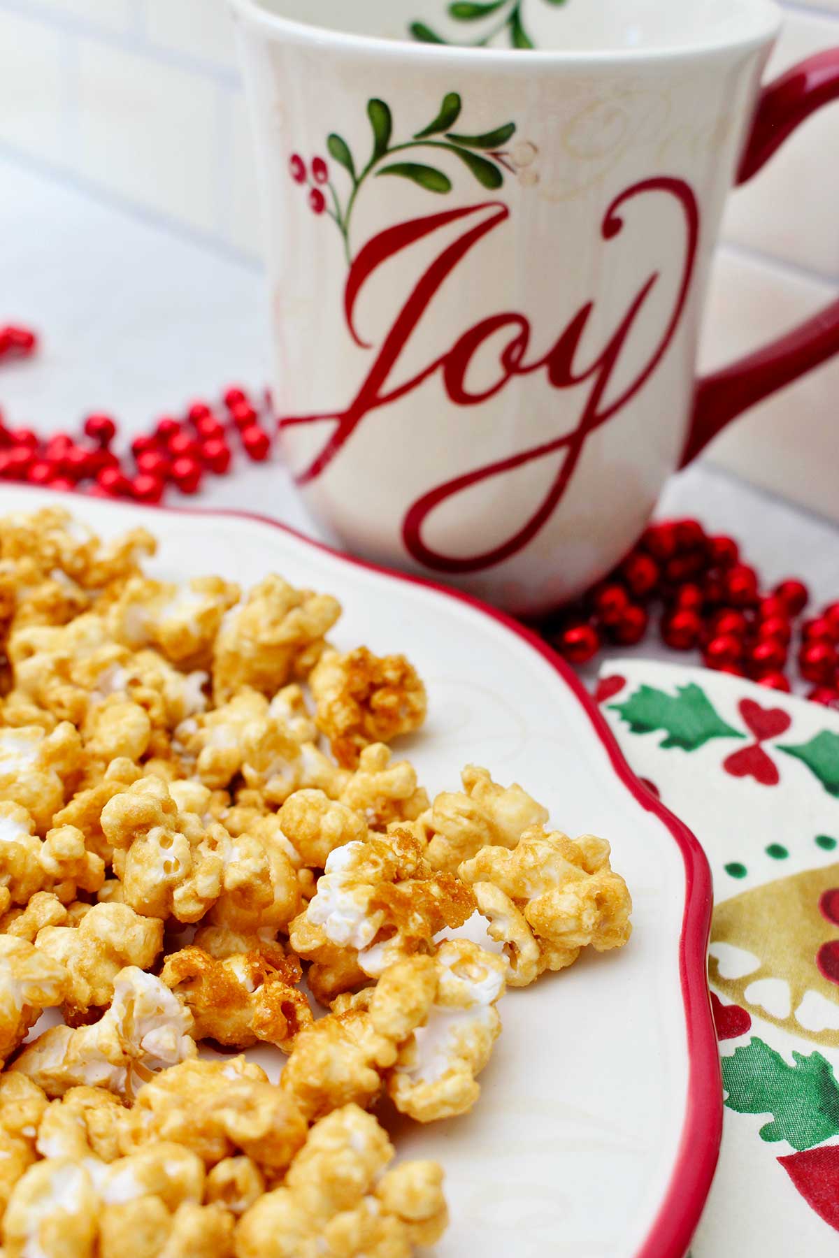 Close up view of caramel corn on a white plate with a red rim, a holiday napkin, red beads and a holiday mug that says "joy".