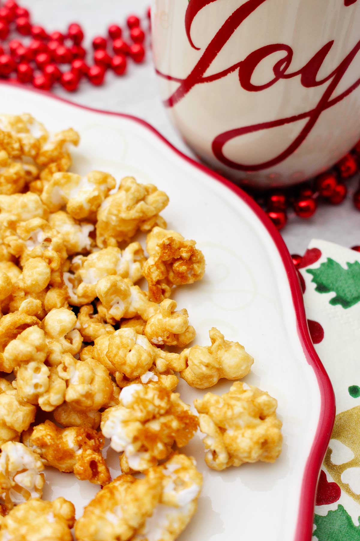 Close up view of caramel corn on a white plate with a red rim, a holiday napkin, red beads and a holiday mug that says "joy".