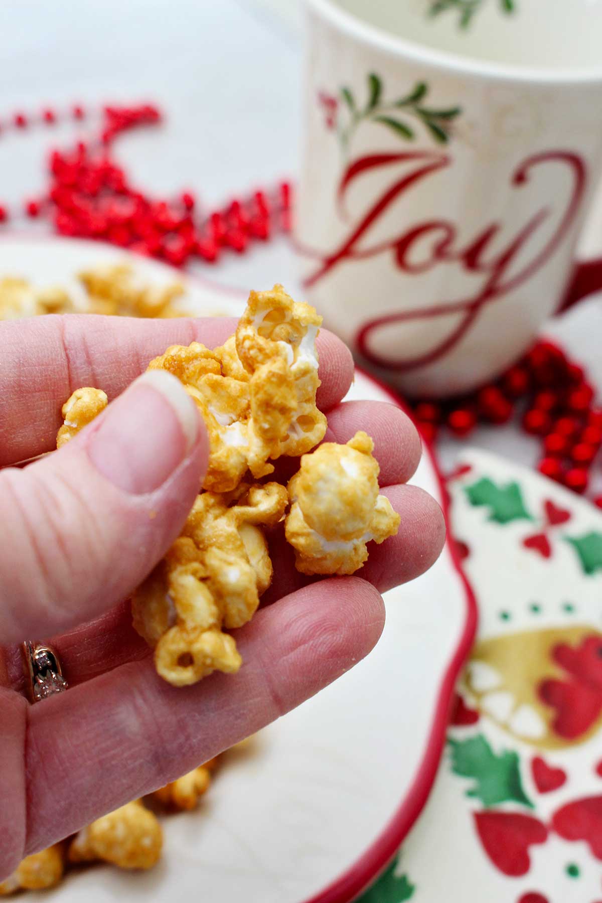 Person holding a handful of caramel corn with a holiday mug and decorations in the background.