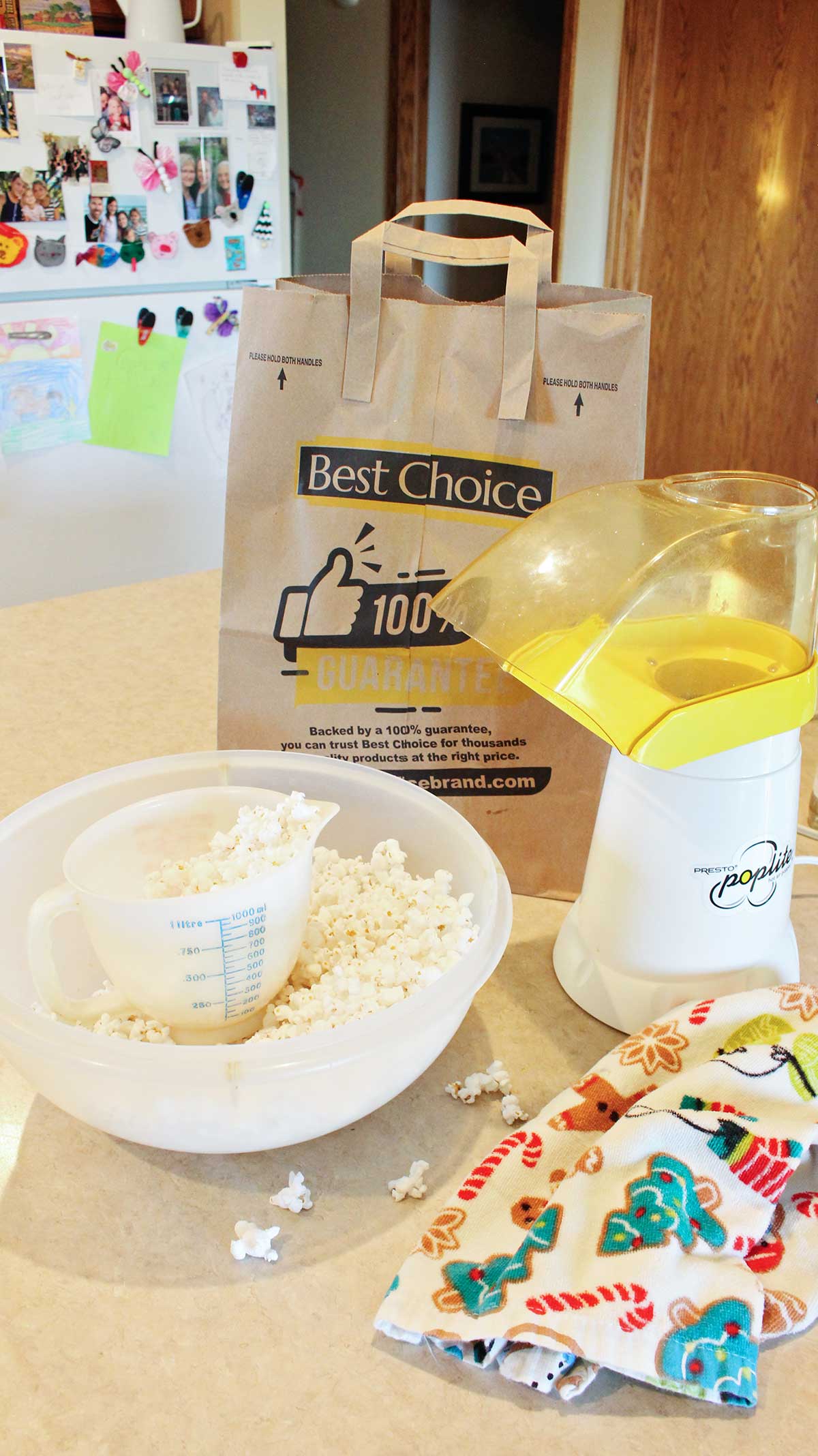 Bowl of popcorn, popcorn maker, holiday linen and paper bag sitting on a kitchen counter.