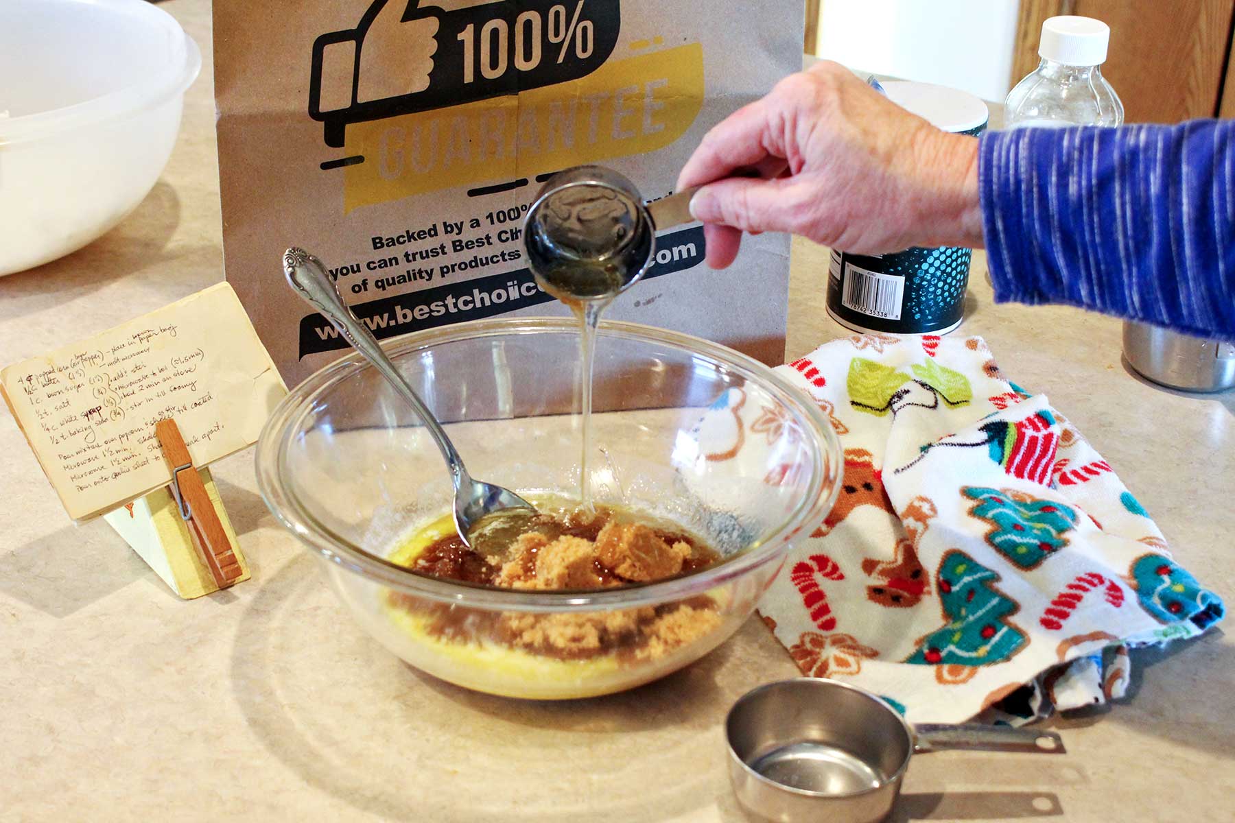 Person pouring corn syrup in a bowl of ingredients for caramel with recipe, linen and other ingredients near by.