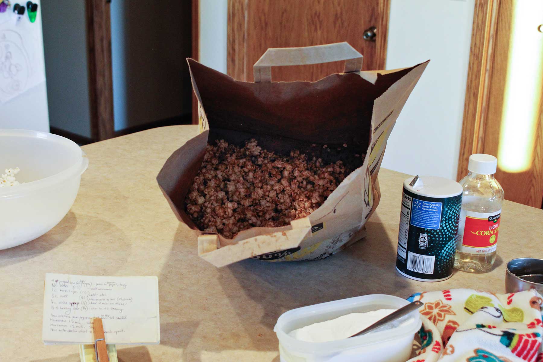 An open paper bag showing caramel corn inside with other supplies sitting on a kitchen counter.