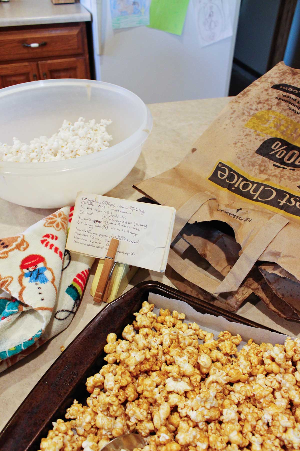 A recipe card holder holding a caramel corn recipe, bowl of popcorn, used paper bag, holiday linen and cookie sheet of caramel corn on a kitchen counter.