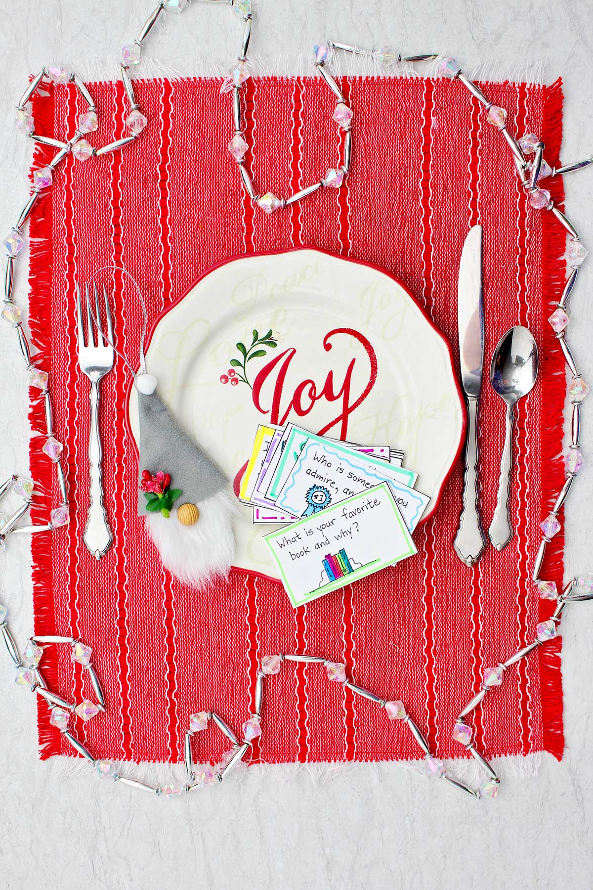 Holiday plate that says "joy" with conversation starter cards on it as well as a gnome decoration sits on a red placemat with place setting. Decorative crystal garland surrounds place setting.