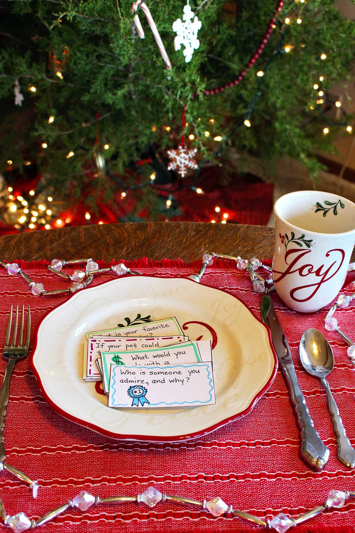 A stack of conversation starter cards resting on a festive holiday plate with a matching mug on a red placemat in front of a Christmas tree.