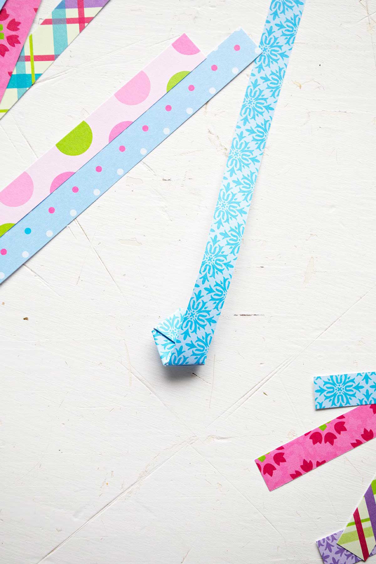 A colorful blue strip of scrapbook paper folded into a pentagon shape, surrounded by strips of paper