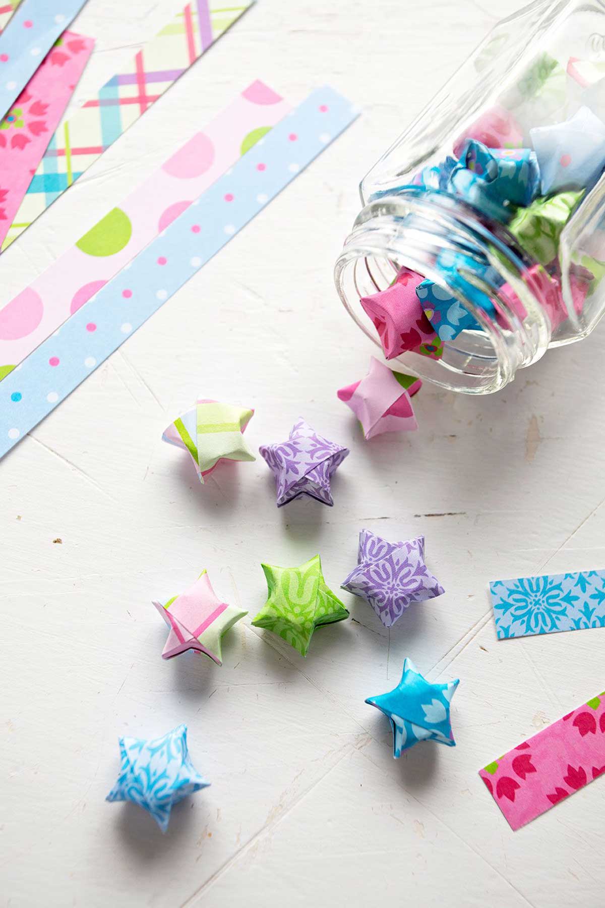 Colorful folded paper stars made from origami or scrapbook paper, falling out of a glass jar