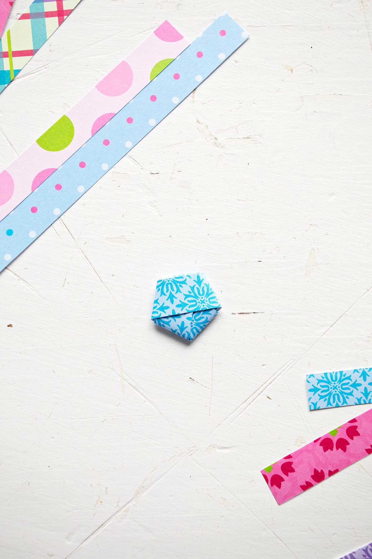 A colorful blue strip of scrapbook paper folded into a pentagon shape, surrounded by strips of paper