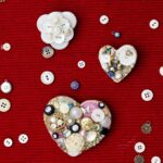 Two heart shaped brooches made from recycled jewelry and buttons with other buttons scattered around sitting on a red background.