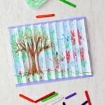 Completed agamograph picture of a tree in two different seasons pasted on a periwinkle paper with crayons, tape and scissors near by.
