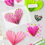 Pink and green paper heart accordions used to decorate cards and valentines with a couple markers and buttons scattered nearby.