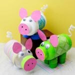 Three DIY piggy banks made from recycled bottles in colors of green, blue and pink sit in front of a yellow backdrop.