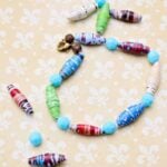Colorful paper beads and blue plastic beads strung together to make a bracelet on a light yellow patterned piece of fabric.