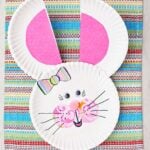 Paper plate bunny with pink ears and cheeks, a pom pom nose, striped bow and google eyes.