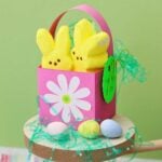 A pink mini Easter basket with a white flower on the center sits on a wooden pedestal filled with candy and Easter grass against a green background.