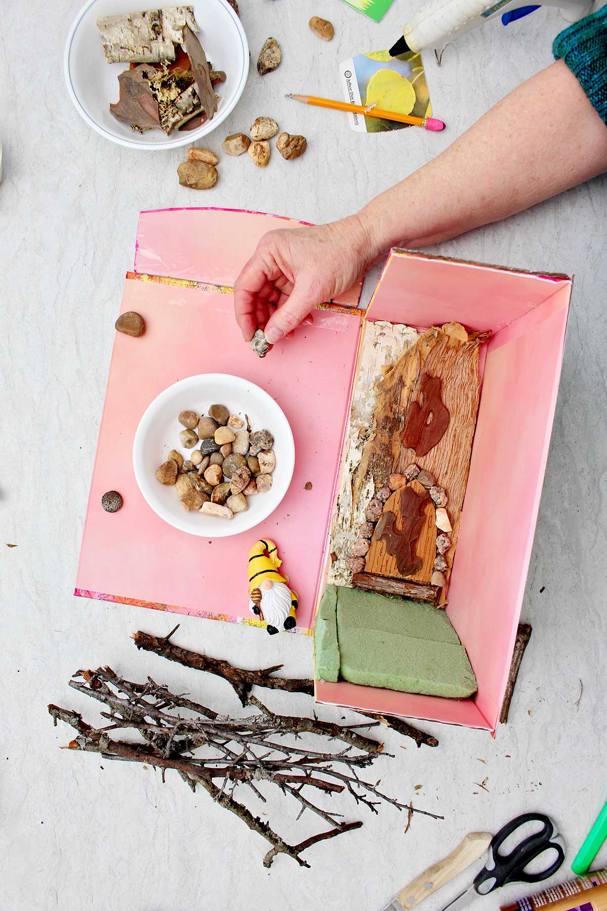 Person gluing small rocks around a doorframe in a miniature forest book nook with other supplies near by.