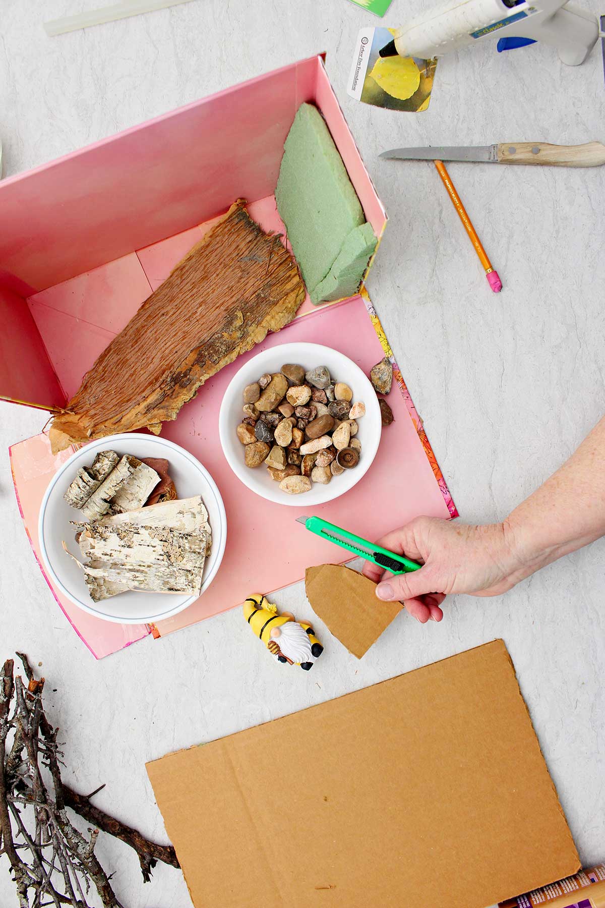 A person holding a rounded door cut out of a piece of cardboard with an exacto knife with other supplies to make a forest book nook near by.