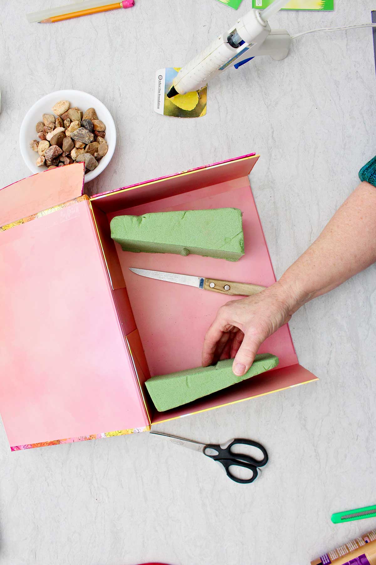 A person placing a cut piece of foam on the bottom of a cut gift box with supplies near by.