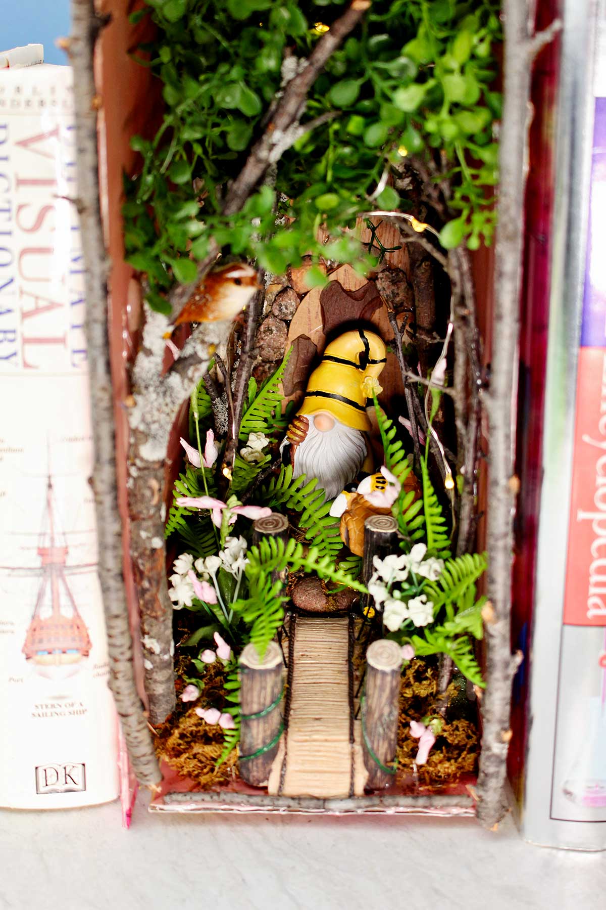 A close up view of a forest book nook with twigs and flowers and a gnome nestled in between old books.