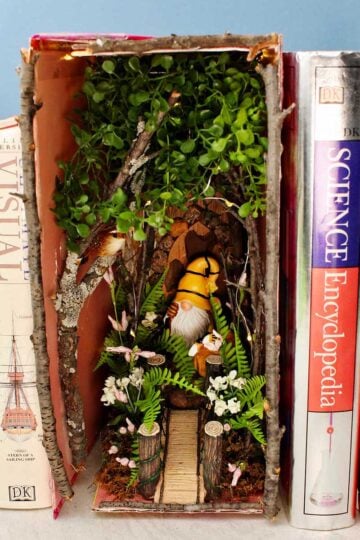 A forest book nook with twigs and flowers and a gnome nestled in between old books.