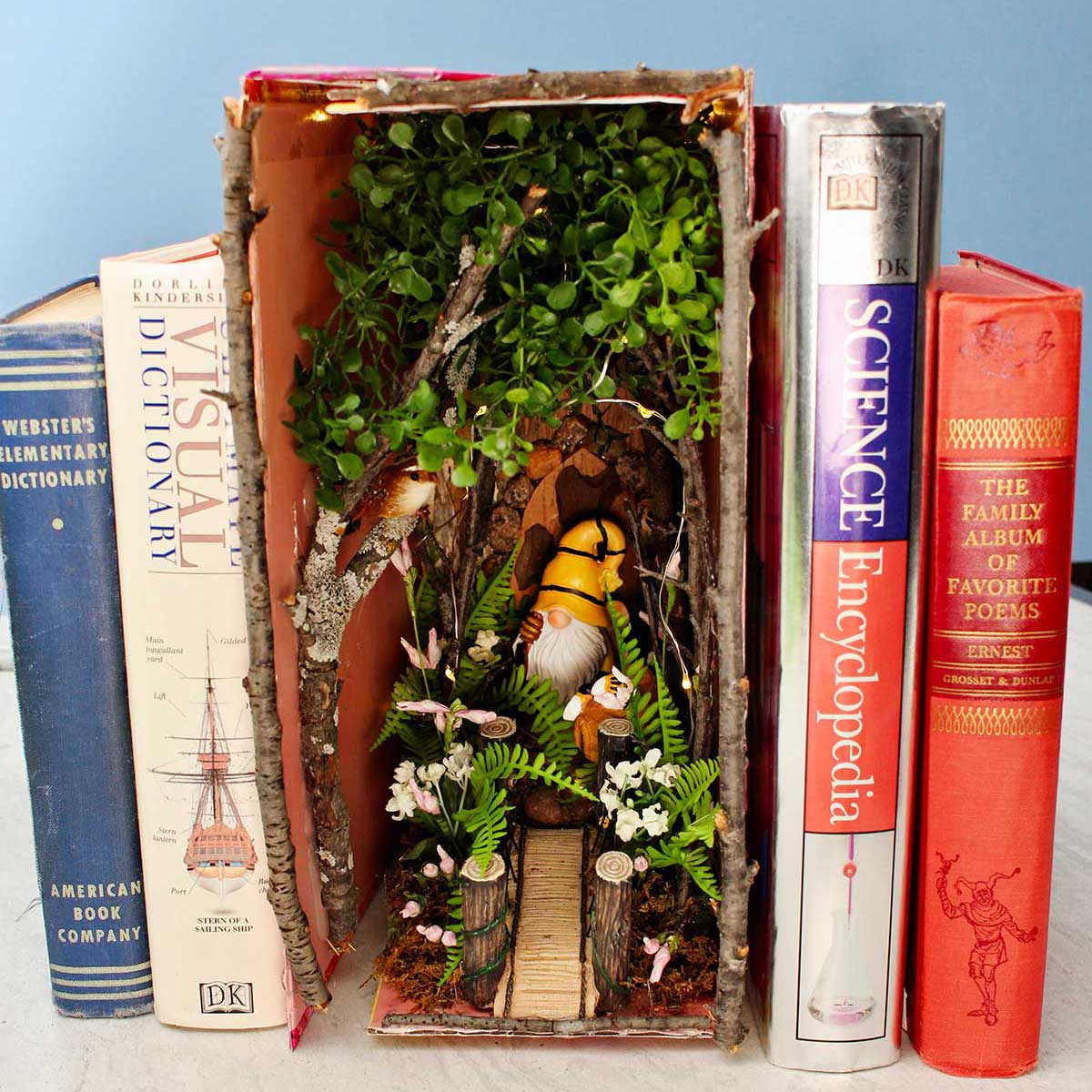 A forest book nook with twigs and flowers and a gnome nestled in between old books.