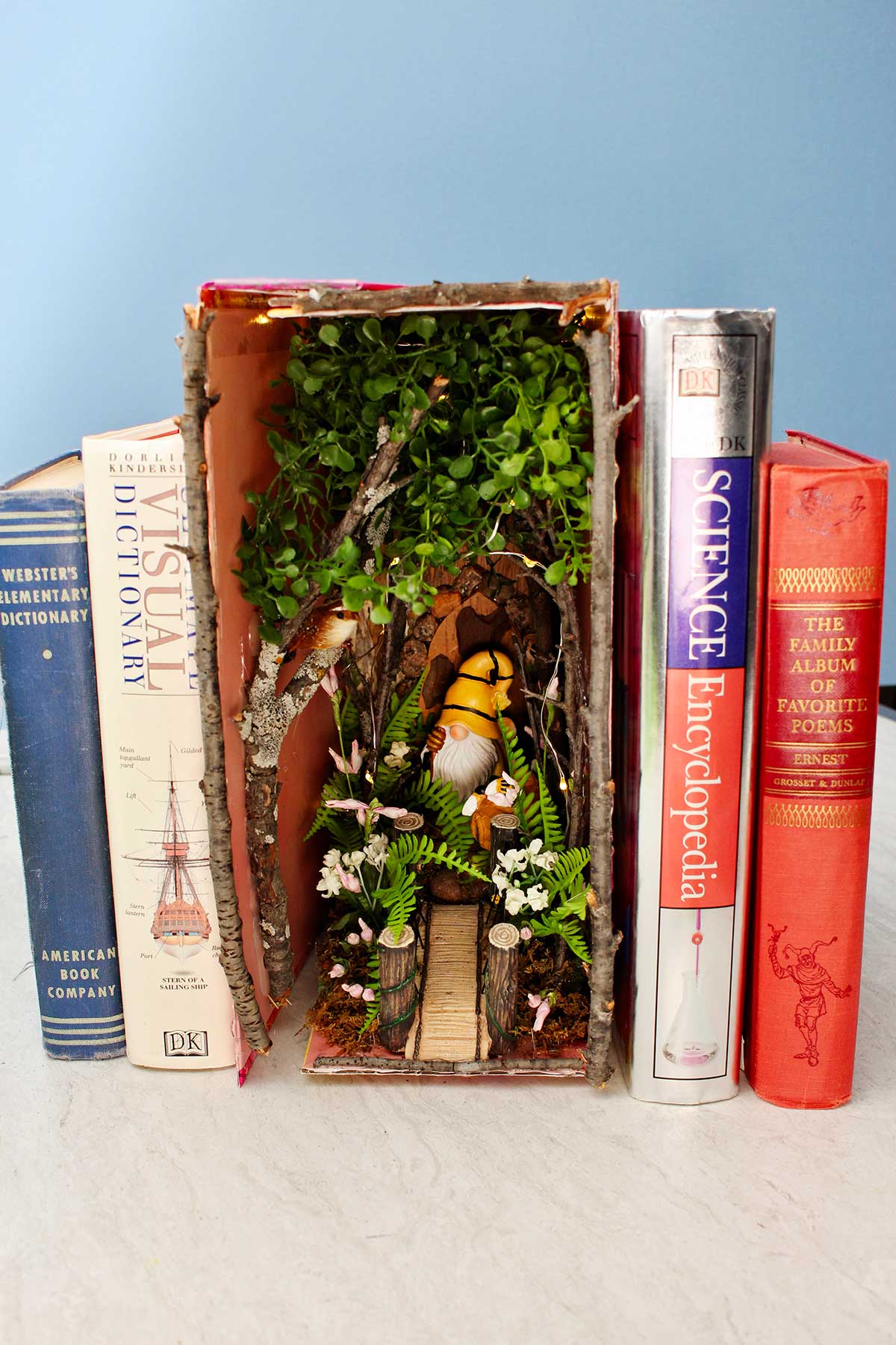 A forest book nook with twigs and flowers and a gnome nestled in between old books.