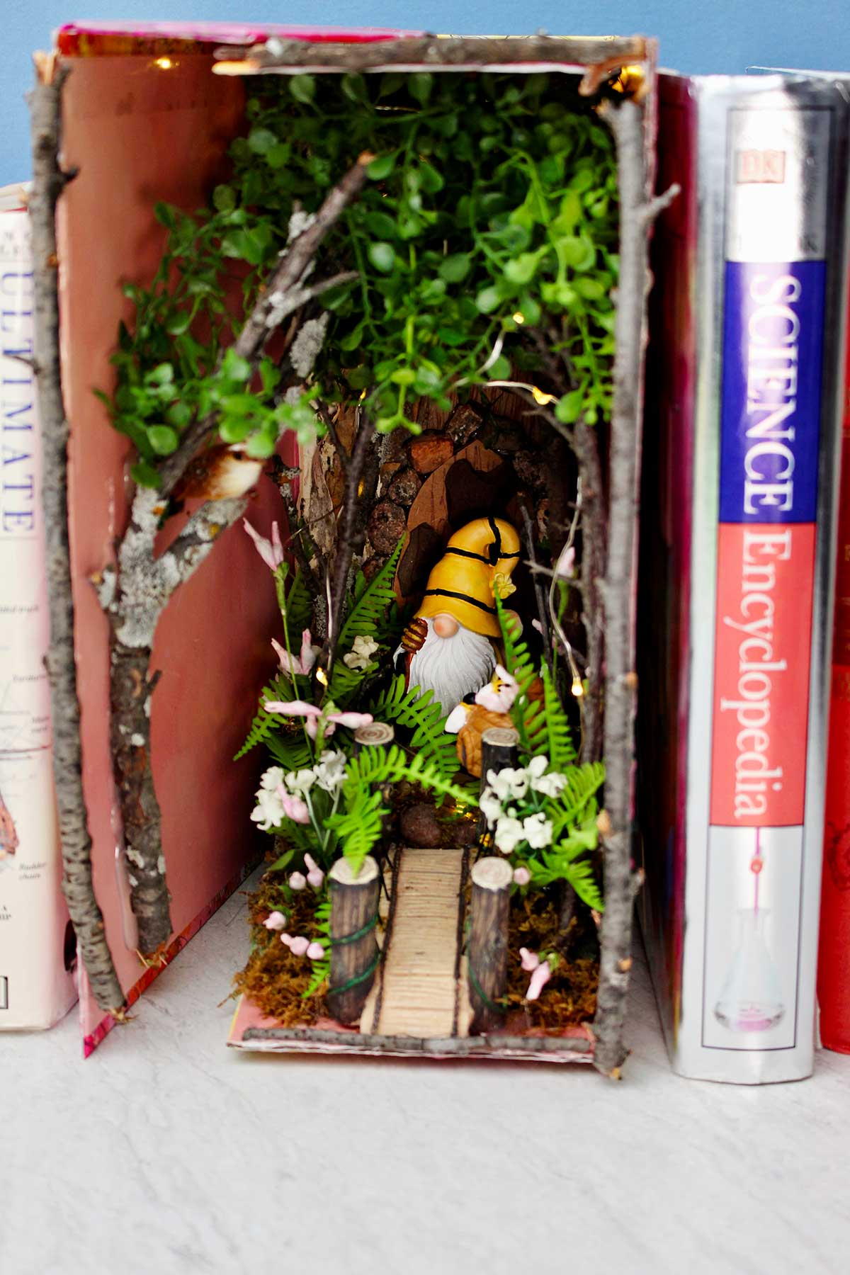 A forest book nook with twigs and flowers and a gnome nestled in between old books.