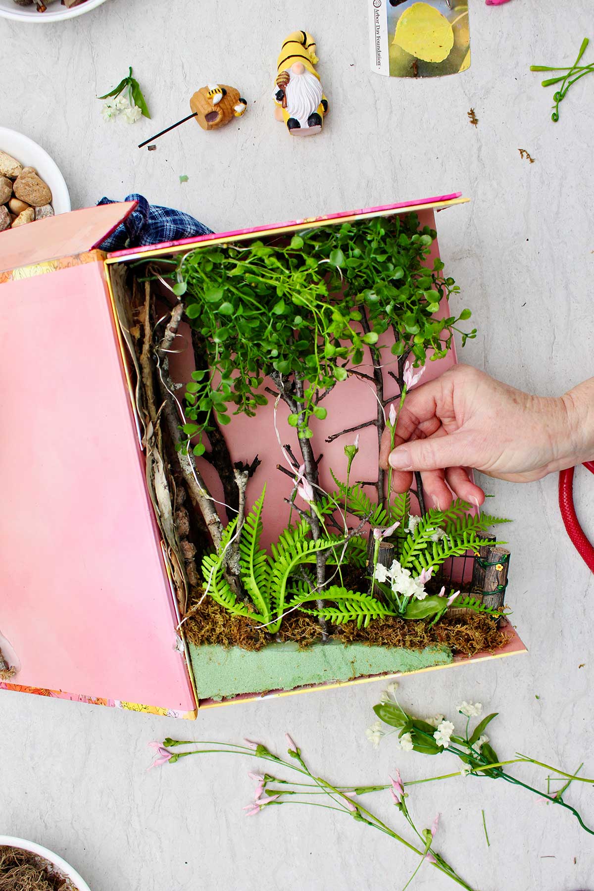 A person placing fake flowers around the miniature forest in the book nook shelf insert.