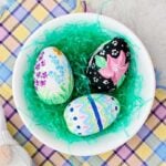 A bowl of three painted wooden Easter eggs in Easter basket grass resting on a pastel plaid cloth.