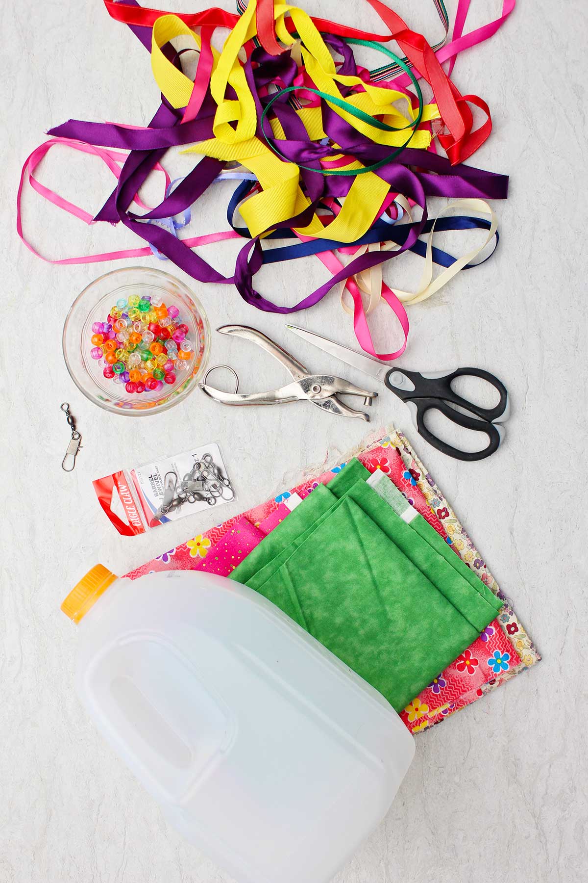 Empty milk jug, a pile of ribbon, stacks of colorful fabric, scissors, a hole punch and beads.