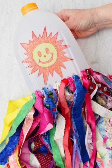 A person. holding a milk jug windsock with a sunshine smiley face on it with many colorful strips of fabric hanging from the bottom.