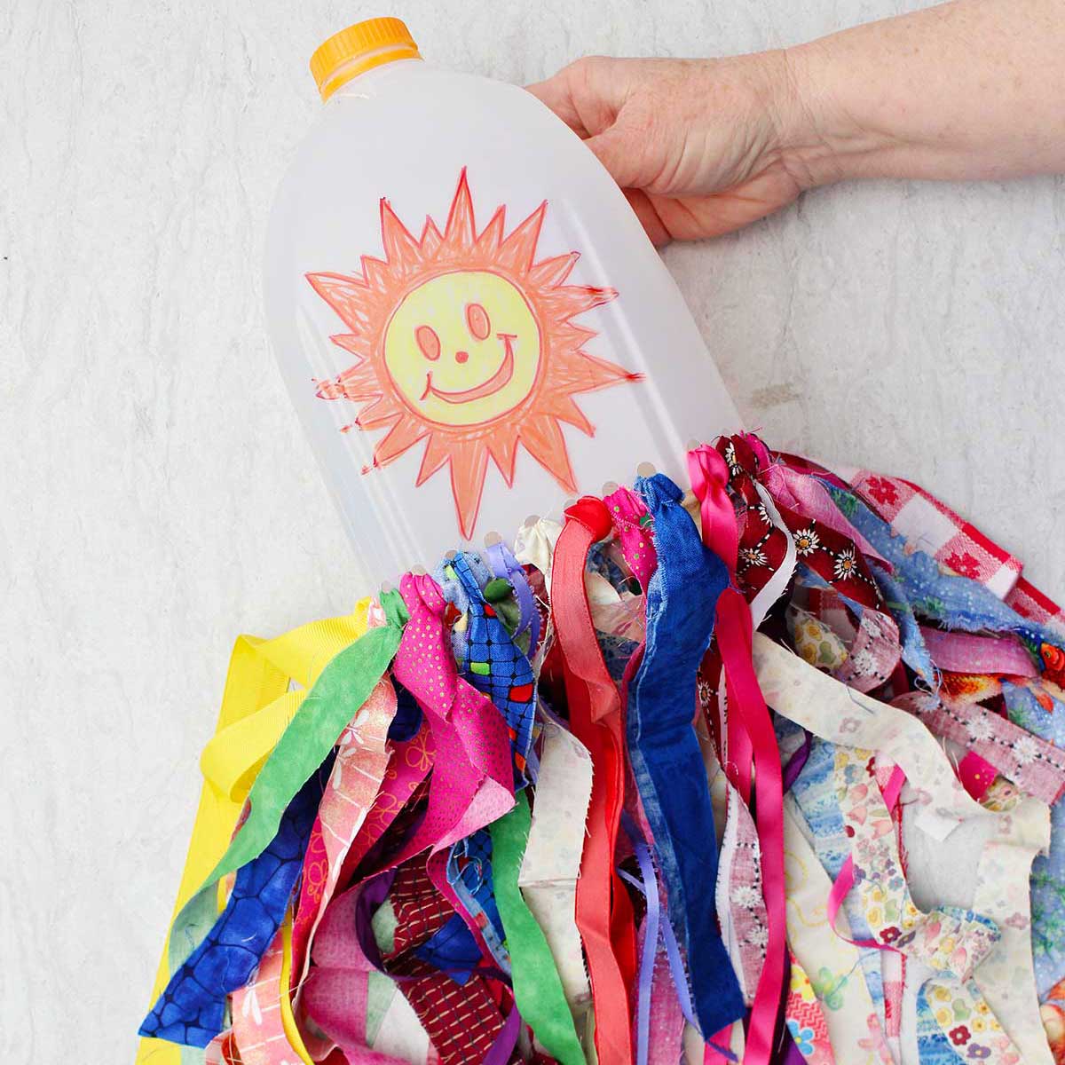A person. holding a milk jug windsock with a sunshine smiley face on it with many colorful strips of fabric hanging from the bottom.