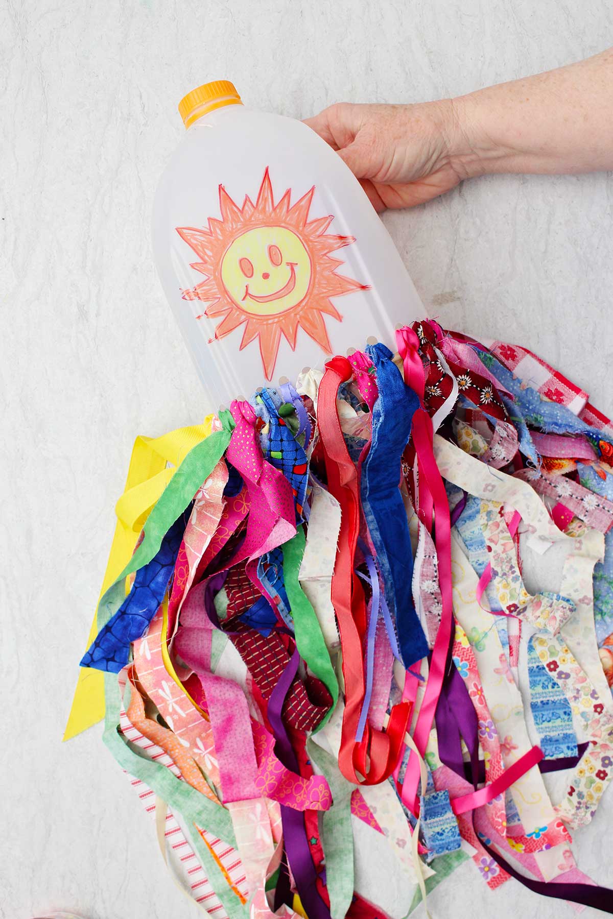 A person. holding a milk jug windsock with a sunshine smiley face on it with many colorful strips of fabric hanging from the bottom.