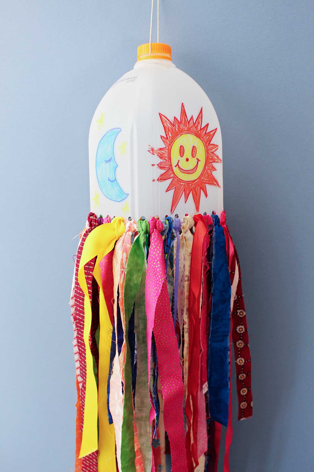 A milk jug windsock with a moon and smiley face sunshine on the jug with many colorful strips of fabric attached to the bottom against a blue backdrop.