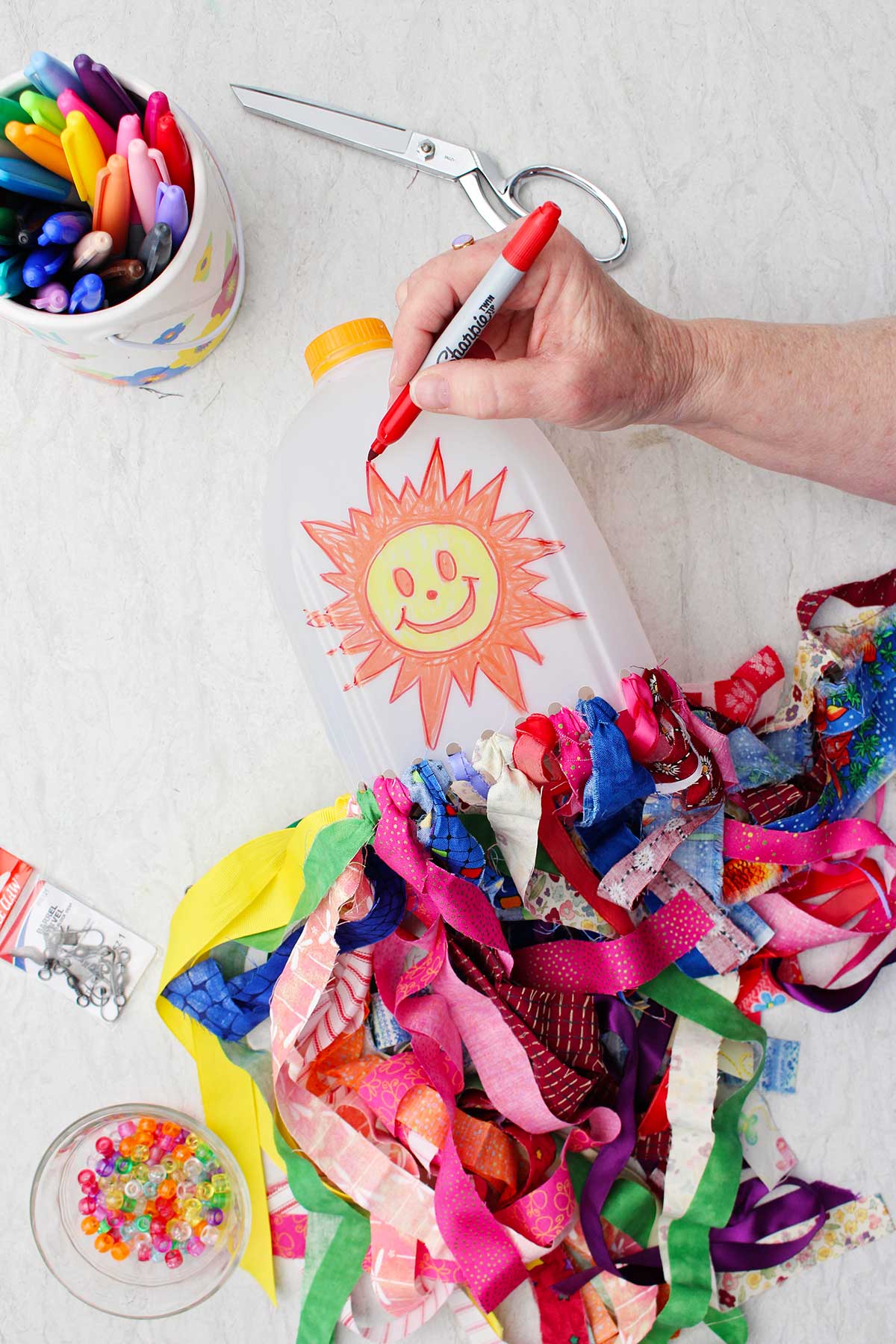 A person outlining a smiley face sunshine drawing on the milk jug windsock with red sharpie.
