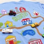 A police car, tractor and yellow Beetle toy cars sitting on a Father's Day T-Shirt painted with a dirt road leading to various buildings.