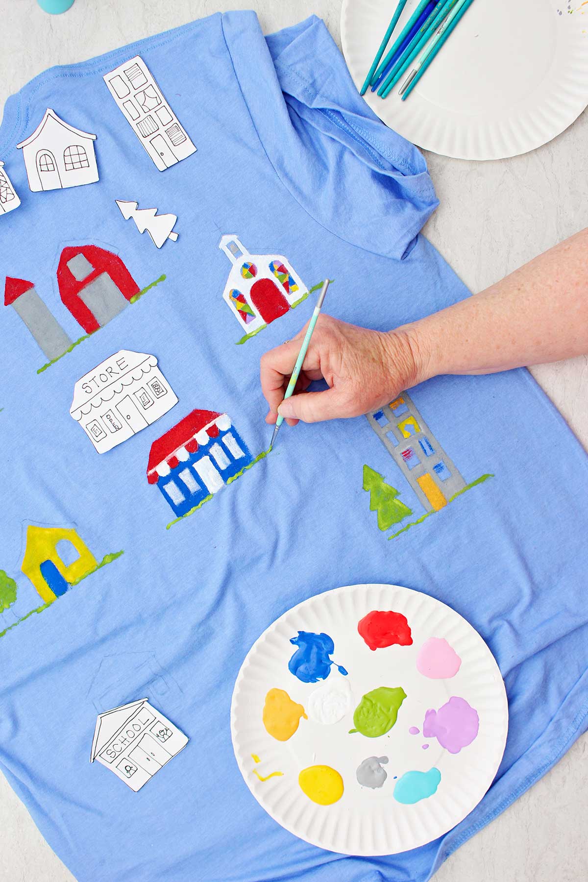 A person painting grass details near a store painted on a blue t-shirt with a plate of paint near by.