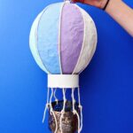 Person holding a completed mini hot air balloon craft against a blue background.