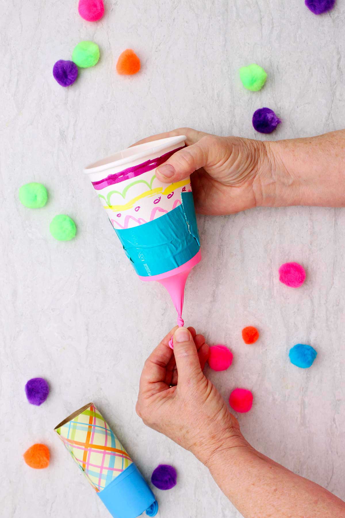 Person pulling the tied end of the balloon on a marshmallow shooter with other pom poms near by.